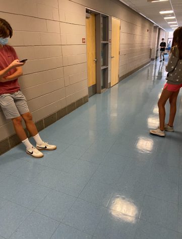 Students waiting in a hallway before their first period classes. On the left side of the hallway, one boy is on his phone and wearing a mas, on the other side there is a girl waiting near her class’ door.