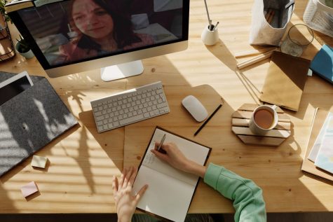 Student studying and taking notes at desk from home, with a Zoom call on a monitor