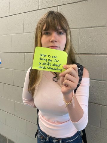 Girl stands against wall with a note that reads., “What is one thing that you like or dislike about block scheduling?”