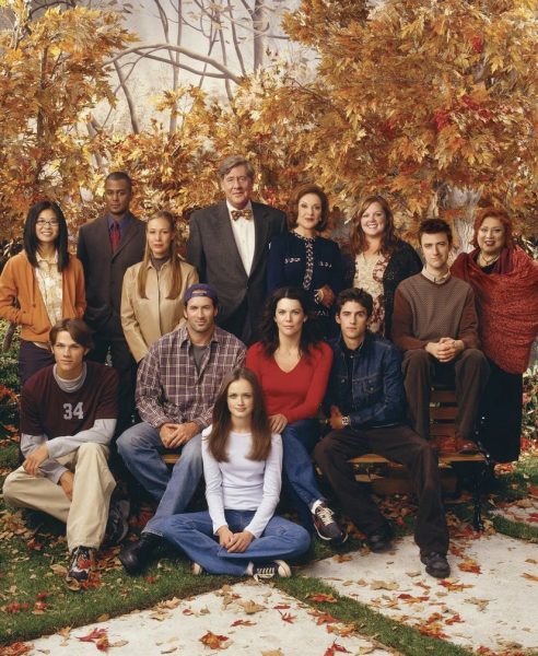 The beloved cast of Gilmore Girls poses in front of a scenic fall backdrop